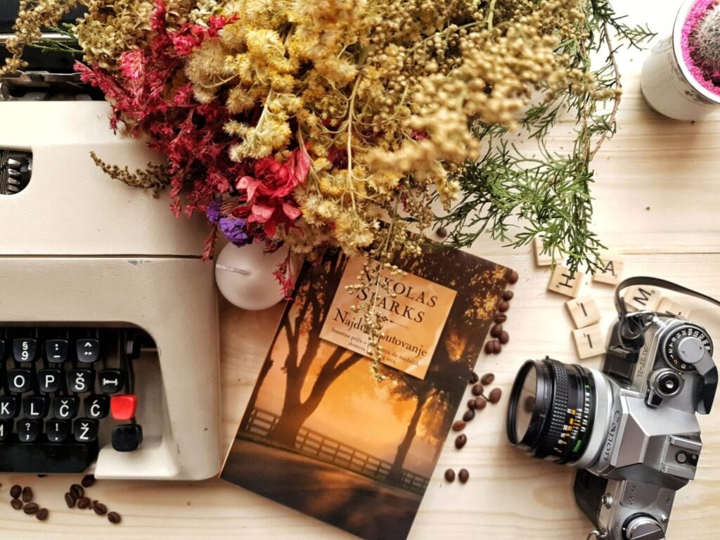 Top view of book and photo camera placed on wooden table near vintage typewriter composed with beautiful dried flowers